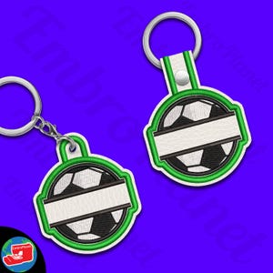 Puede incluir: Dos llaveros de balón de fútbol. Cada llavero presenta un diseño de balón de fútbol con una pancarta blanca para personalización, delineada en verde. Los llaveros tienen un llavero plateado y un respaldo de polipiel blanco.
