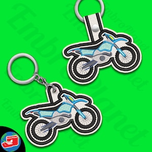Puede incluir: Dos llaveros de moto de cross azul y blanco. Cada llavero presenta un diseño detallado de una moto de cross con ruedas negras y un cuadro azul. Los llaveros están unidos a llaveros de plata.
