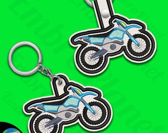 Op de afbeelding: Twee blauwe en witte dirt bike sleutelhangers. Elke sleutelhanger heeft een gedetailleerd ontwerp van een dirt bike met zwarte wielen en een blauw frame. De sleutelhangers zijn bevestigd aan zilveren sleutelringen.