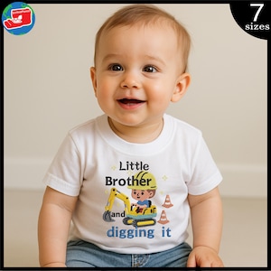 以下が含まれることがあります： 白いTシャツを着た笑顔の赤ちゃん。カラフルなグラフィックが特徴です。グラフィックは、黄色の掘削機を操作する漫画の建設作業員で、「Little Brother and digging it」というテキストが表示されています。