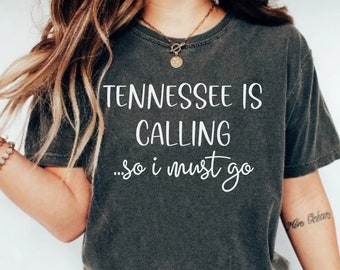 以下が含まれることがあります： 「TENNESSEE IS CALLING ...so i must go」の白文字がプリントされた濃いグレーのTシャツ。クルーネックと半袖のデザインです。快適でカジュアルなスタイルです。