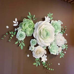 Puede incluir: Una decoraci&oacute;n de pared hecha de flores de papel en tonos de blanco y verde. Las flores est&aacute;n dispuestas en un racimo e incluyen rosas, peon&iacute;as y otros tipos de flores. Las flores est&aacute;n fijadas a una pared marr&oacute;n.