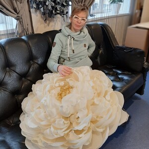 Pu&ograve; includere: Un grande fiore di carta bianco che ricorda una peonia, seduto su un divano in pelle nera. Una persona &egrave; seduta sul divano dietro il fiore.