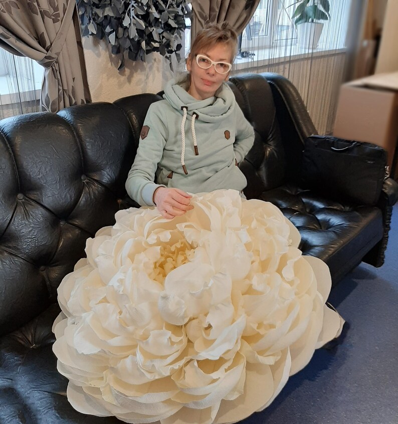 Pu&ograve; includere: Un grande fiore di carta bianco che ricorda una peonia, seduto su un divano in pelle nera. Una persona &egrave; seduta sul divano dietro il fiore.