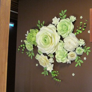 Puede incluir: Una decoraci&oacute;n de pared hecha de flores de papel. Las flores son blancas y verdes y est&aacute;n dispuestas en un racimo. Las flores est&aacute;n fijadas a una pared marr&oacute;n.
