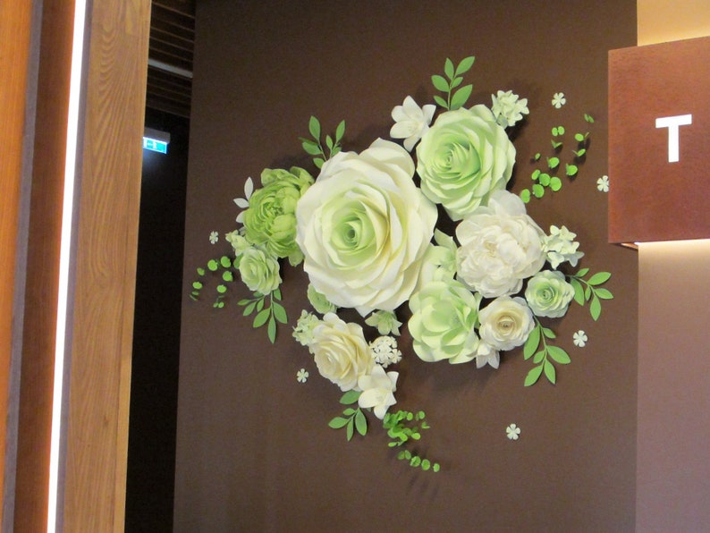 Puede incluir: Una decoraci&oacute;n de pared hecha de flores de papel. Las flores son blancas y verdes y est&aacute;n dispuestas en un racimo. Las flores est&aacute;n fijadas a una pared marr&oacute;n.