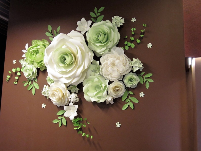 Puede incluir: Una decoraci&oacute;n de pared con un grupo de flores de papel en tonos blancos y verdes. Las flores est&aacute;n dispuestas en cascada, con la flor m&aacute;s grande en el centro. Las flores est&aacute;n hechas de papel y tienen un aspecto realista.