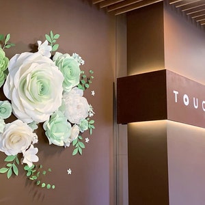 Puede incluir: Una pared marr&oacute;n con una gran disposici&oacute;n de flores de papel en tonos blancos y verdes. Las flores est&aacute;n dispuestas en forma de semic&iacute;rculo. La pared tambi&eacute;n tiene un letrero que dice "TOUCH DAY SPA".