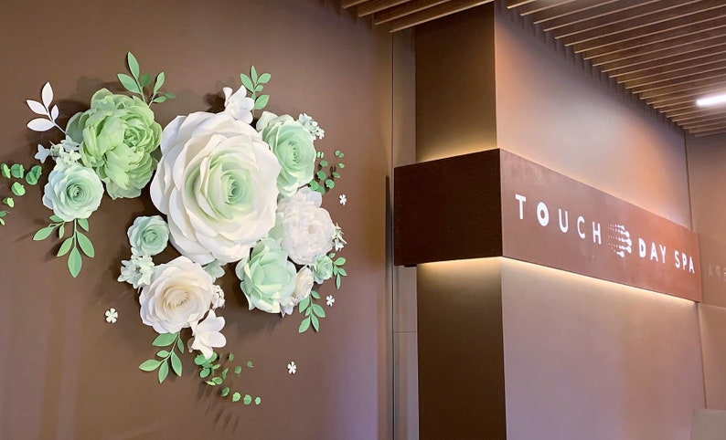 Puede incluir: Una pared marr&oacute;n con una gran disposici&oacute;n de flores de papel en tonos blancos y verdes. Las flores est&aacute;n dispuestas en forma de semic&iacute;rculo. La pared tambi&eacute;n tiene un letrero que dice "TOUCH DAY SPA".