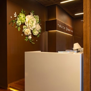 Puede incluir: Un mostrador de recepci&oacute;n blanco con una pared de madera marr&oacute;n detr&aacute;s. La pared tiene una disposici&oacute;n de flores de papel y un letrero que dice "TOUCH DAY SPA".