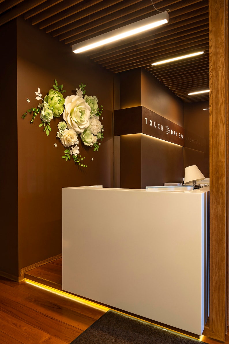 Puede incluir: Un mostrador de recepci&oacute;n blanco con una pared de madera marr&oacute;n detr&aacute;s. La pared tiene una disposici&oacute;n de flores de papel y un letrero que dice "TOUCH DAY SPA".