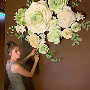 Puede incluir: Una mujer est&aacute; decorando una pared con grandes flores de papel. Las flores son de color blanco y verde y est&aacute;n dispuestas en un racimo. La pared es de color marr&oacute;n oscuro.