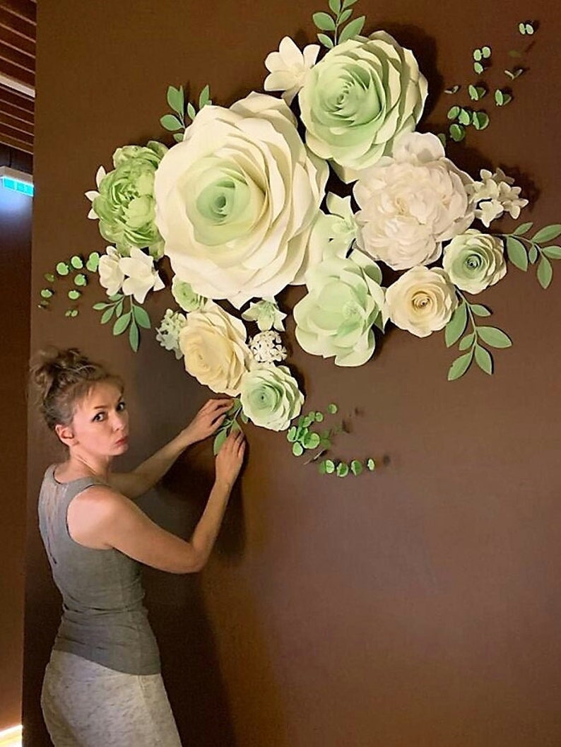 Puede incluir: Una mujer est&aacute; decorando una pared con grandes flores de papel. Las flores son de color blanco y verde y est&aacute;n dispuestas en un racimo. La pared es de color marr&oacute;n oscuro.