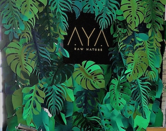 May include: A green paper backdrop with a large, black sign that reads "AYA RAW NATURE". The backdrop is made of paper leaves in various shades of green.