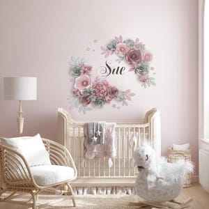 K&ouml;nnte beinhalten: Ein rosa-graues Blumen-Wandtattoo f&uuml;r das Kinderzimmer mit dem Namen "Sue" in kursivschrift. Das Wandtattoo befindet sich &uuml;ber einem wei&szlig;en Kinderbett mit einer rosa-grauen Decke.