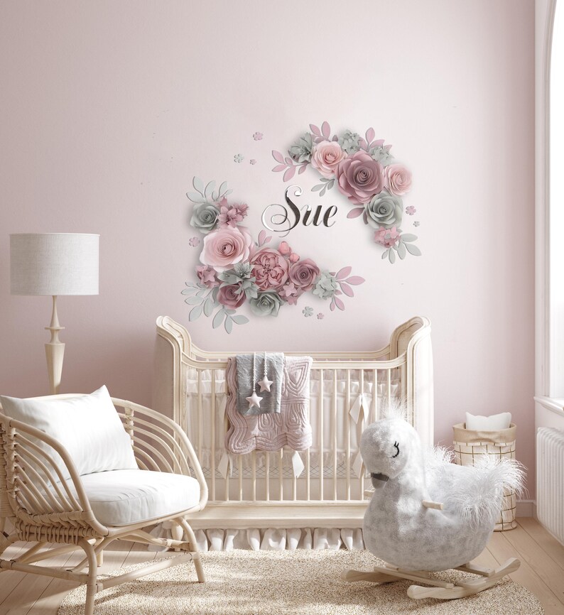 K&ouml;nnte beinhalten: Ein rosa-graues Blumen-Wandtattoo f&uuml;r das Kinderzimmer mit dem Namen "Sue" in kursivschrift. Das Wandtattoo befindet sich &uuml;ber einem wei&szlig;en Kinderbett mit einer rosa-grauen Decke.