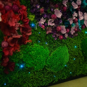 May include: A close-up of a green moss wall with dried flowers and small blue lights. The moss is arranged in a pattern of mounds and the flowers are in shades of pink, purple, and red.