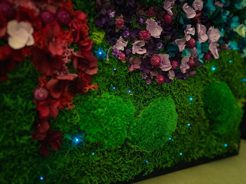 May include: A close-up of a green moss wall with dried flowers and small blue lights. The moss is arranged in a pattern of mounds and the flowers are in shades of pink, purple, and red.