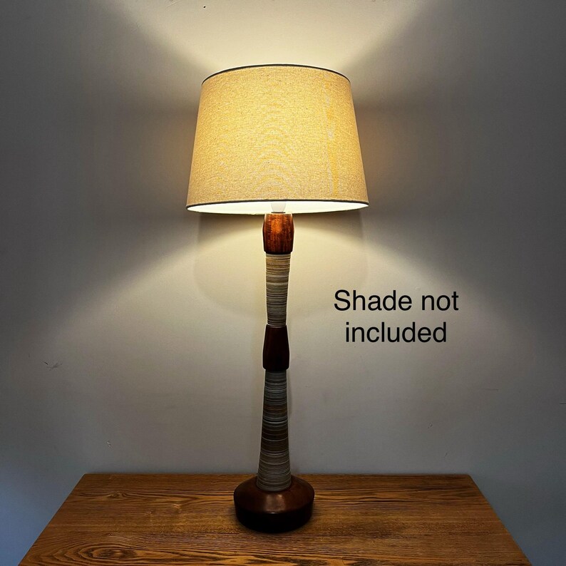 May include: A tall table lamp with a dark brown wooden base and a slender, multi-textured stem featuring alternating bands of dark wood and grey-toned material. The lamp is topped with a beige fabric lampshade. Text on the right reads: Shade not included.