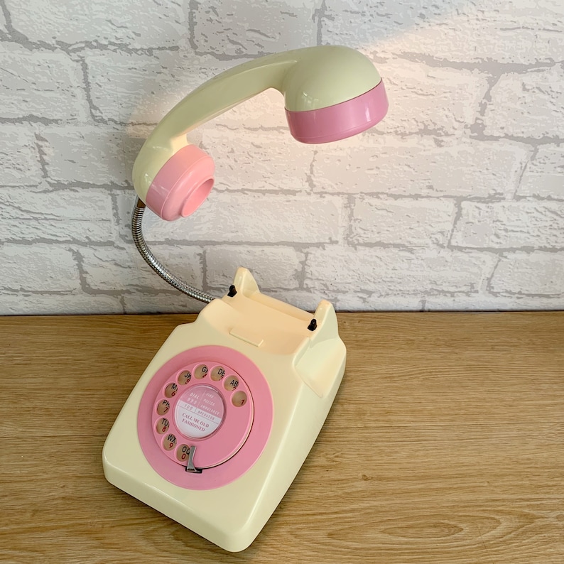 May include: A vintage-style rotary phone with a pink and cream color scheme. The phone has a flexible arm that holds a pink and cream lamp. The phone has a dial with the text "Call me old fashioned".