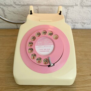 May include: A vintage rotary dial phone with a cream-colored body and a pink rotary dial. The dial has black letters and numbers, and the phone has a black cord. The phone has a label that reads "Call me old fashioned" with the words "Fire, Police, Ambulance, Operator" listed above it.