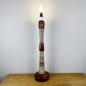 May include: A tall, slender table lamp featuring a stacked design of polished dark brown wood segments and textured, multi-colored striped sections. The lamp base is circular and wooden, topped with a single, glowing white candle-shaped light bulb.