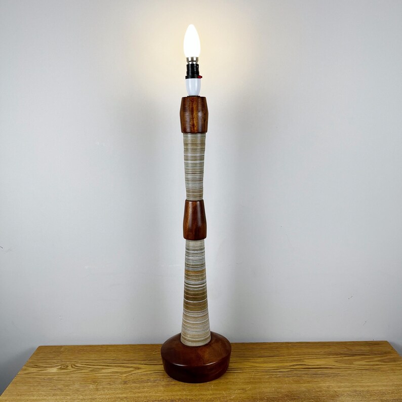 May include: A tall, slender table lamp featuring a stacked design of polished dark brown wood segments and textured, multi-colored striped sections. The lamp base is circular and wooden, topped with a single, glowing white candle-shaped light bulb.