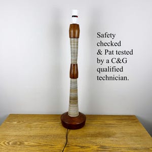 May include: A tall table lamp base featuring a cylindrical body with alternating sections of dark brown wood and light-colored, horizontally striped material. The text reads: Safety checked & Pat tested by a C&G qualified technician.