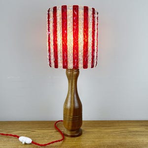 May include: A table lamp featuring a cylindrical shade made of alternating vertical red and clear textured glass strips. The lamp has a smooth, turned wooden base with a tapered neck and a red fabric-covered power cord with a white inline switch.