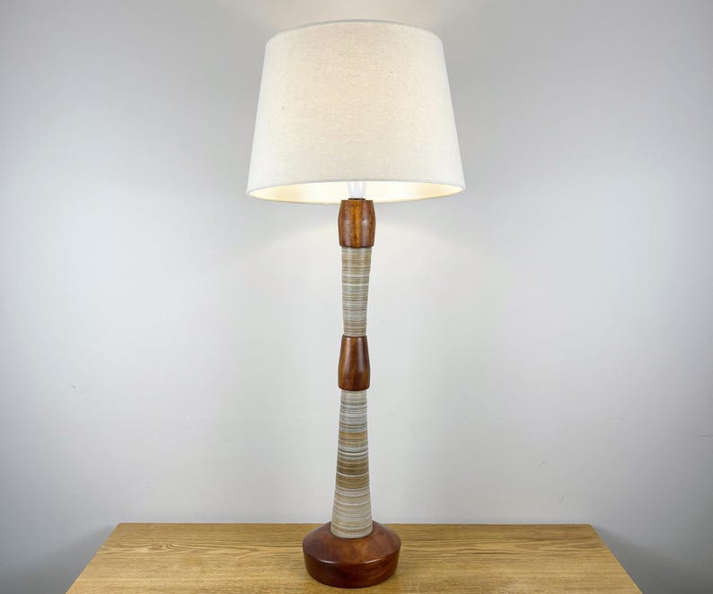 May include: A table lamp featuring a tall, slender base composed of alternating sections of polished dark wood and a textured, horizontally striped ceramic or stone material, topped with a simple, off-white, tapered fabric drum lampshade.