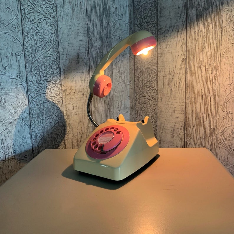 May include: A vintage-style rotary phone lamp with a pink and cream color scheme. The phone has a rotary dial and a pink lampshade. The lamp is on a table with a light wood finish.