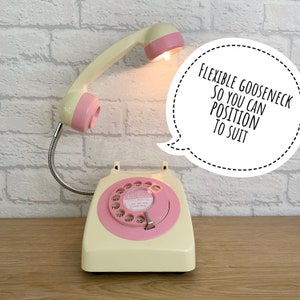 May include: A vintage-style rotary phone with a pink and cream color scheme. The phone has a flexible gooseneck lamp attached to the handset, allowing for adjustable lighting. The lamp is turned on and illuminating the phone's dial. The text on the dial reads "CALL MELB FASININED". The phone is on a wooden surface. The text "FLEXIBLE GOOSENECK SO YOU CAN POSITION TO SUIT" is in a speech bubble above the phone.