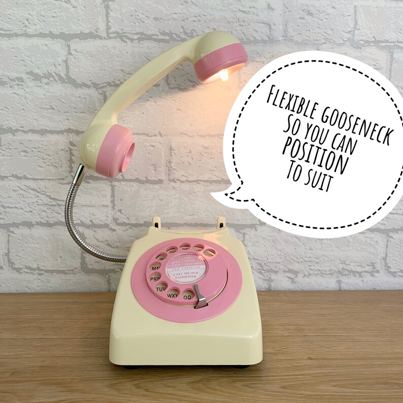 May include: A vintage-style rotary phone with a pink and cream color scheme. The phone has a flexible gooseneck lamp attached to the handset, allowing for adjustable lighting. The lamp is turned on and illuminating the phone's dial. The text on the dial reads "CALL MELB FASININED". The phone is on a wooden surface. The text "FLEXIBLE GOOSENECK SO YOU CAN POSITION TO SUIT" is in a speech bubble above the phone.