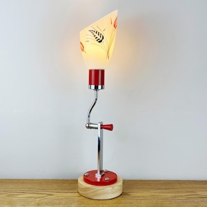 May include: A table lamp featuring a conical glass shade with a black and red leaf pattern. The lamp has a chrome-plated metal arm, a red cylindrical base, and a light-colored wooden circular stand. A red handle is attached to the arm.