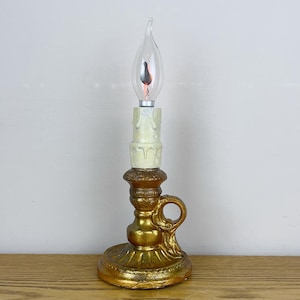 May include: A vintage-style electric candle lamp with a gold-colored base and handle. The lamp features a cream-colored candle-shaped section and a clear, flame-shaped light bulb. The base has ornate detailing.