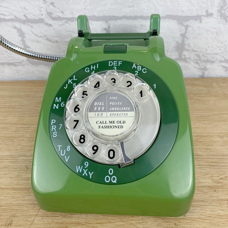 May include: A green rotary dial phone with a silver cord. The phone has a white dial with black numbers and letters. The dial has the text "FIRE POLICE AMBULANCE 999" and "100 OPERATOR" printed on it. The phone also has the text "CALL ME OLD FASHIONED" printed on it.