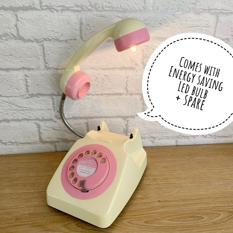 May include: A vintage-style rotary phone with a pink dial and a cream-colored body. The phone has a pink and cream-colored handset that doubles as a lamp with a light bulb. The text "COMES WITH ENERGY SAVING LED BULB + SPARE" is in a speech bubble.