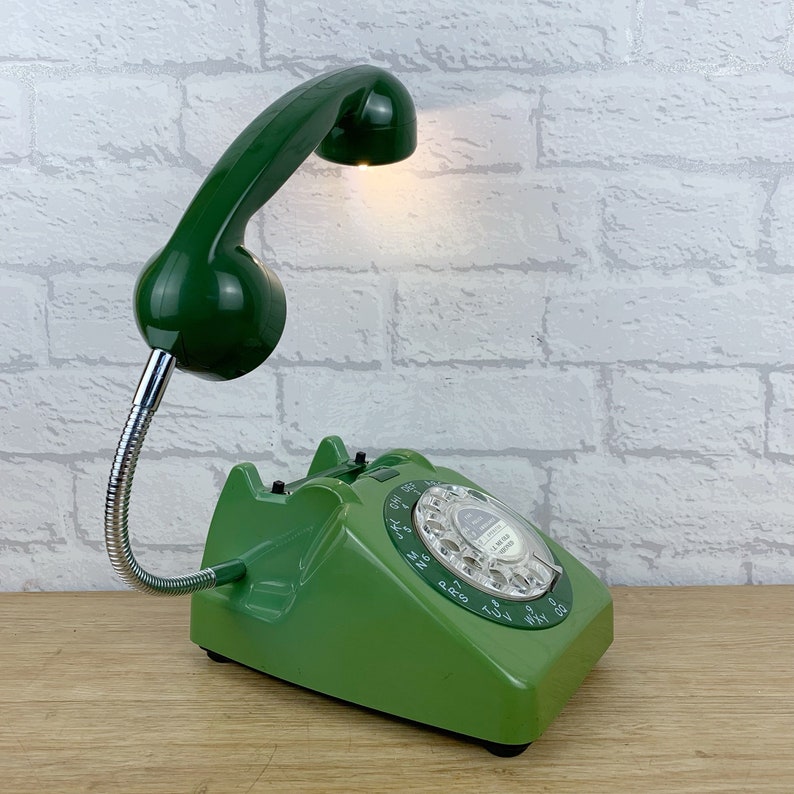 May include: A green rotary dial phone with a green lamp attached to the handset. The lamp has a flexible arm and a round shade. The phone has a white dial with black numbers and a white button.