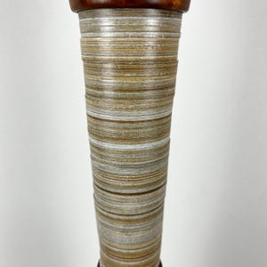 May include: A tall, tapered cylindrical object featuring a stacked, layered appearance with horizontal bands in shades of tan, beige, white, and brown. The top section is finished with a smooth, dark reddish-brown polished wood cap.