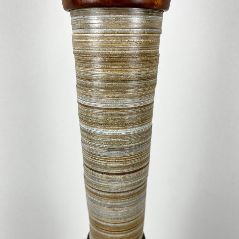 May include: A tall, tapered cylindrical object featuring a stacked, layered appearance with horizontal bands in shades of tan, beige, white, and brown. The top section is finished with a smooth, dark reddish-brown polished wood cap.