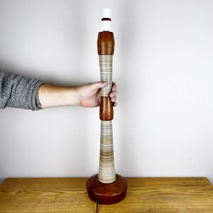 May include: A tall table lamp base featuring a cylindrical design with alternating sections of polished dark brown wood and light-colored, horizontally layered material. The lamp has a white socket at the top and a wide, circular wooden base.