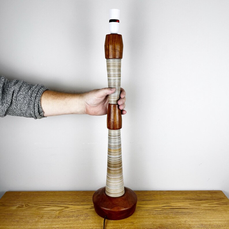 May include: A tall table lamp base featuring a cylindrical design with alternating sections of polished dark brown wood and light-colored, horizontally layered material. The lamp has a white socket at the top and a wide, circular wooden base.