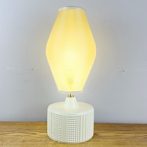 May include: A cream-coloured table lamp with a textured base and a ribbed, hourglass-shaped shade. The shade emits a warm, yellow glow. The lamp has a gold-coloured accent at the base of the shade. The lamp is on a wooden surface.