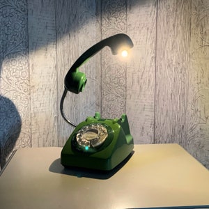 May include: A green rotary phone with a built-in reading lamp. The phone has a green dial and a green handset. The lamp is attached to the handset and has a white light bulb.
