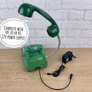 May include: Green rotary phone lamp with a flexible arm and a 12V power supply. The phone has a green rotary dial and a green handset. The lamp is on and the handset is pointing up. The power supply is black and has a UK plug.