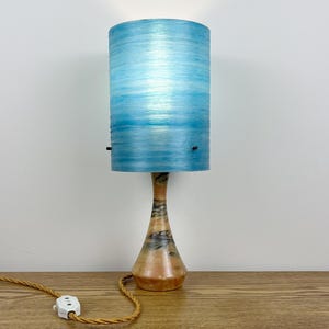 May include: A table lamp featuring a cylindrical light blue shade with horizontal textured lines and a tapered, smooth wooden base with natural dark grain patterns. The lamp includes a tan fabric-covered electrical cord with a white plastic switch.