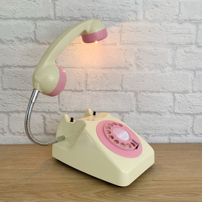 May include: A vintage rotary phone lamp with a cream-colored base and a pink rotary dial. The phone's handset is attached to a flexible arm and has a pink light bulb at the end.