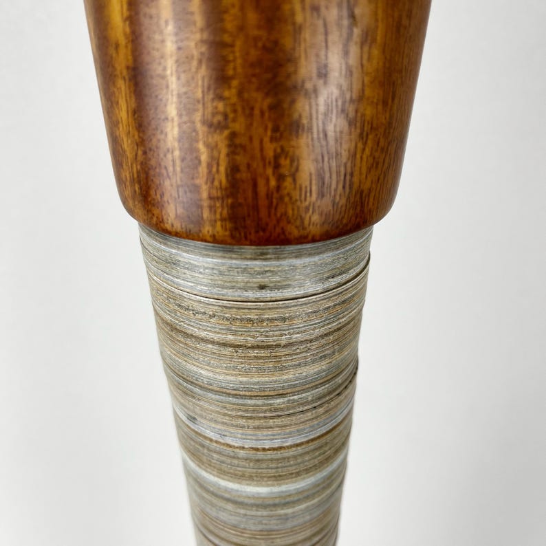 May include: A cylindrical object featuring a smooth, polished dark brown wooden top section with visible grain patterns, transitioning into a tapered base wrapped in numerous thin, light gray and silver metallic horizontal bands.