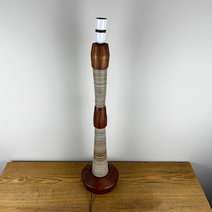 May include: A tall table lamp base featuring a stacked design of dark brown wood sections and cylindrical segments with fine, horizontal grey and white striped patterns. The lamp has a white socket at the top and a circular wooden base.