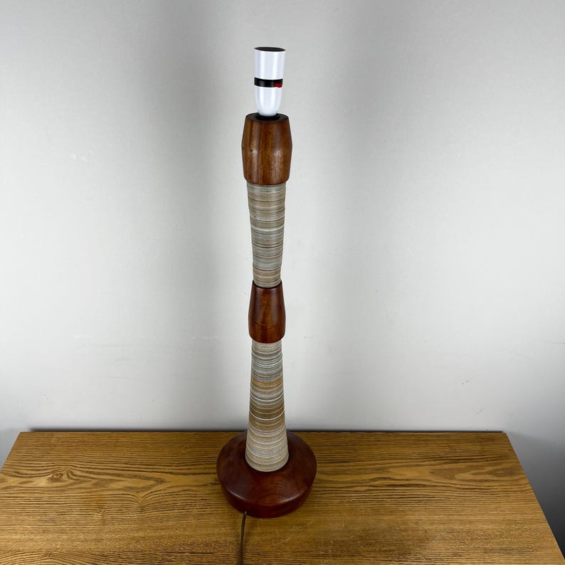 May include: A tall table lamp base featuring a stacked design of dark brown wood sections and cylindrical segments with fine, horizontal grey and white striped patterns. The lamp has a white socket at the top and a circular wooden base.